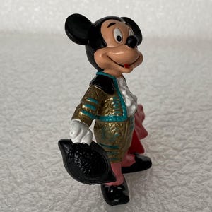 May include: A small Mickey Mouse figurine dressed in a gold and teal matador outfit with a white ruffled shirt. The character holds a black hat in one hand and has a red cape draped behind, featuring black ears and a painted face with a smile.