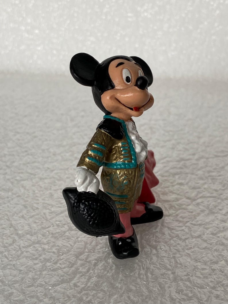May include: A small Mickey Mouse figurine dressed in a gold and teal matador outfit with a white ruffled shirt. The character holds a black hat in one hand and has a red cape draped behind, featuring black ears and a painted face with a smile.