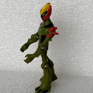 May include: An articulated action figure featuring a green, plant-like body with four arms, red shoulder spikes, and a yellow and red head crest. The figure has a black chest emblem and visible silver joints at the shoulders, elbows, and knees.