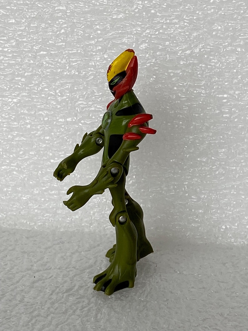 May include: An articulated action figure featuring a green, plant-like body with four arms, red shoulder spikes, and a yellow and red head crest. The figure has a black chest emblem and visible silver joints at the shoulders, elbows, and knees.