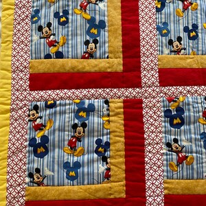Pode incluir: Um edredom colorido com um padr&atilde;o repetido do Mickey Mouse. O edredom tem um fundo listrado azul e branco com figuras do Mickey Mouse em vermelho, amarelo e preto. O edredom &eacute; bordado com tecido vermelho, dourado e branco.