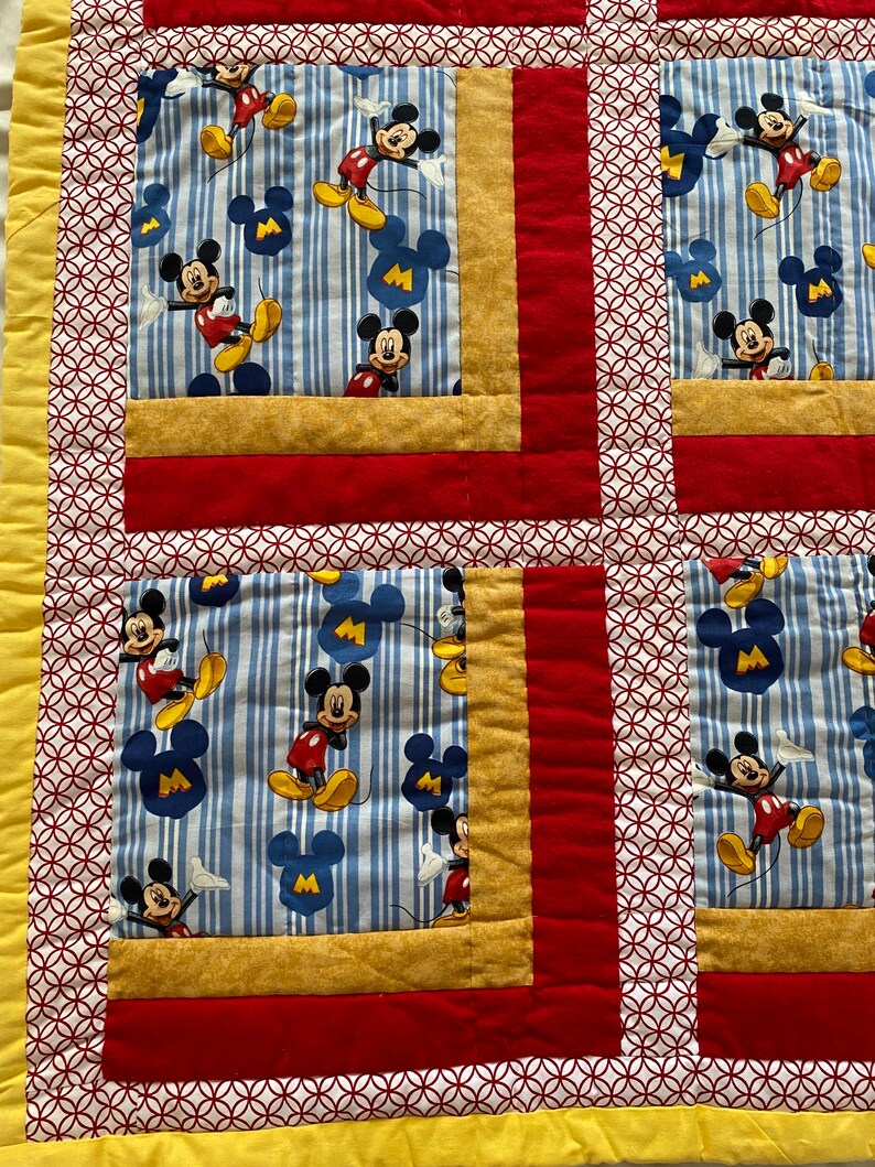 Pode incluir: Um edredom colorido com um padr&atilde;o repetido do Mickey Mouse. O edredom tem um fundo listrado azul e branco com figuras do Mickey Mouse em vermelho, amarelo e preto. O edredom &eacute; bordado com tecido vermelho, dourado e branco.