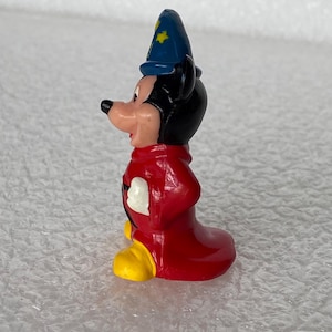 Pu&ograve; includere: Una piccola statuetta in plastica stampata raffigurante un personaggio di topo dei cartoni animati che indossa un cappello da mago blu con stelle gialle, una lunga tunica rossa e grandi scarpe gialle. Il personaggio &egrave; mostrato di profilo.