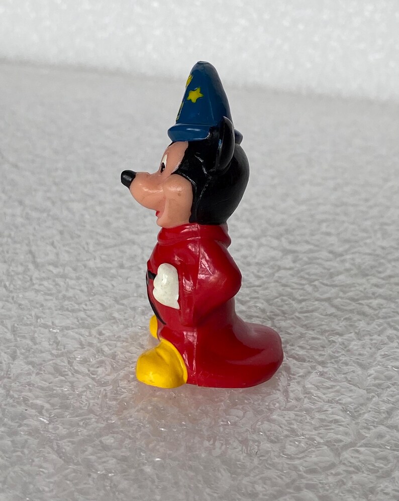 Pu&ograve; includere: Una piccola statuetta in plastica stampata raffigurante un personaggio di topo dei cartoni animati che indossa un cappello da mago blu con stelle gialle, una lunga tunica rossa e grandi scarpe gialle. Il personaggio &egrave; mostrato di profilo.