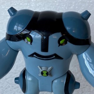 May include: A light blue action figure with black accents and green eyes. The figure has a distinctive head shape, large arms, and a symbol on its chest. The figure is a toy.