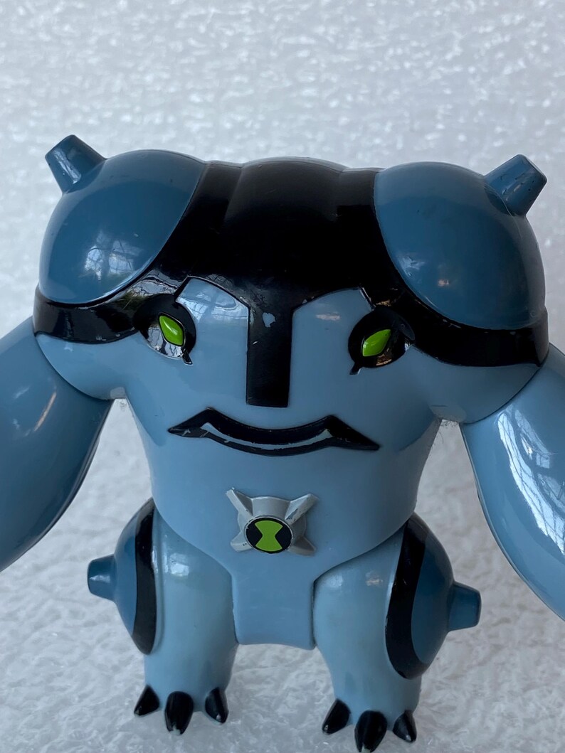 May include: A light blue action figure with black accents and green eyes. The figure has a distinctive head shape, large arms, and a symbol on its chest. The figure is a toy.
