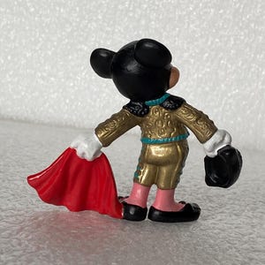 May include: A small figurine of a cartoon mouse dressed as a bullfighter, wearing a gold jacket with teal trim, pink pants, and black shoes. The figure holds a bright red cape in one hand and a black hat in the other, viewed from the back.