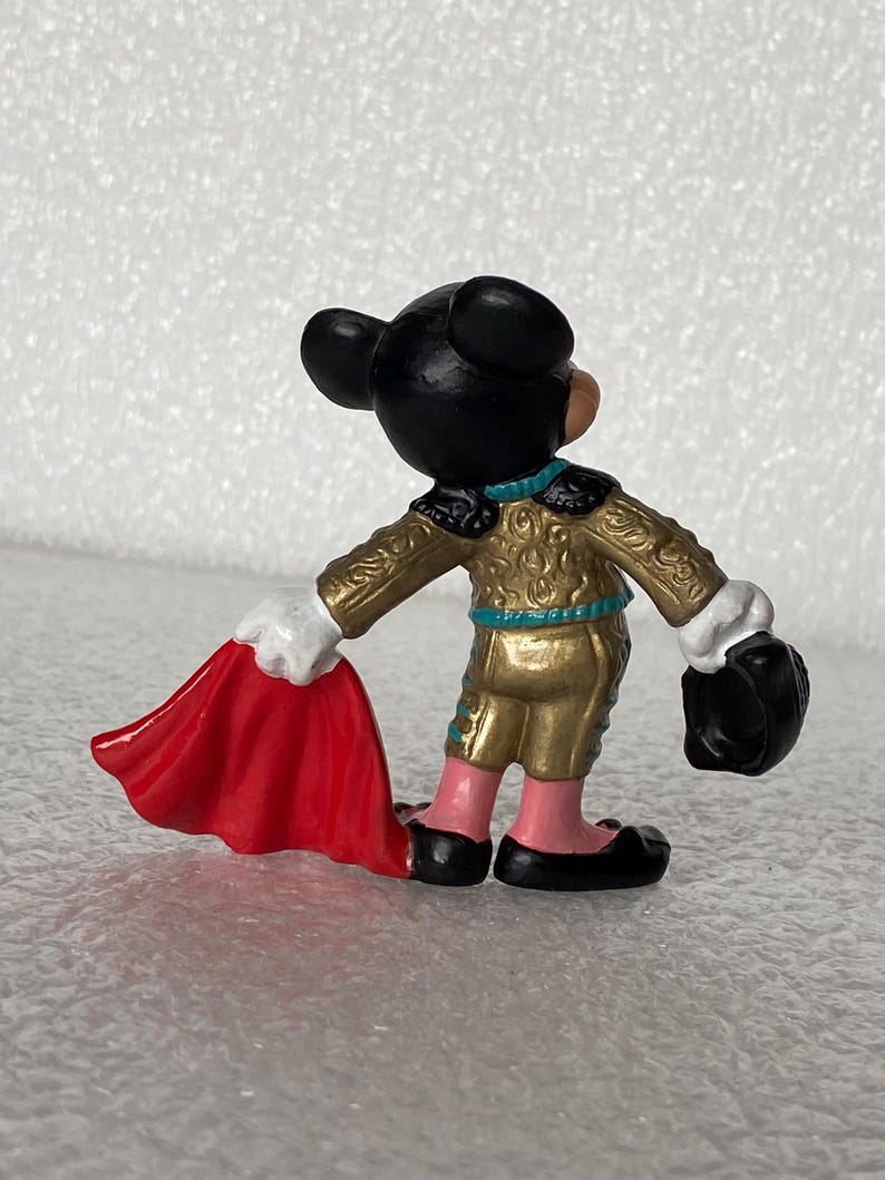 May include: A small figurine of a cartoon mouse dressed as a bullfighter, wearing a gold jacket with teal trim, pink pants, and black shoes. The figure holds a bright red cape in one hand and a black hat in the other, viewed from the back.