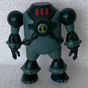 May include: A teal robot action figure with a rounded head and three red rectangles. It features large spherical shoulders, articulated limbs, and a green and black emblem on its chest. The figure is a toy.
