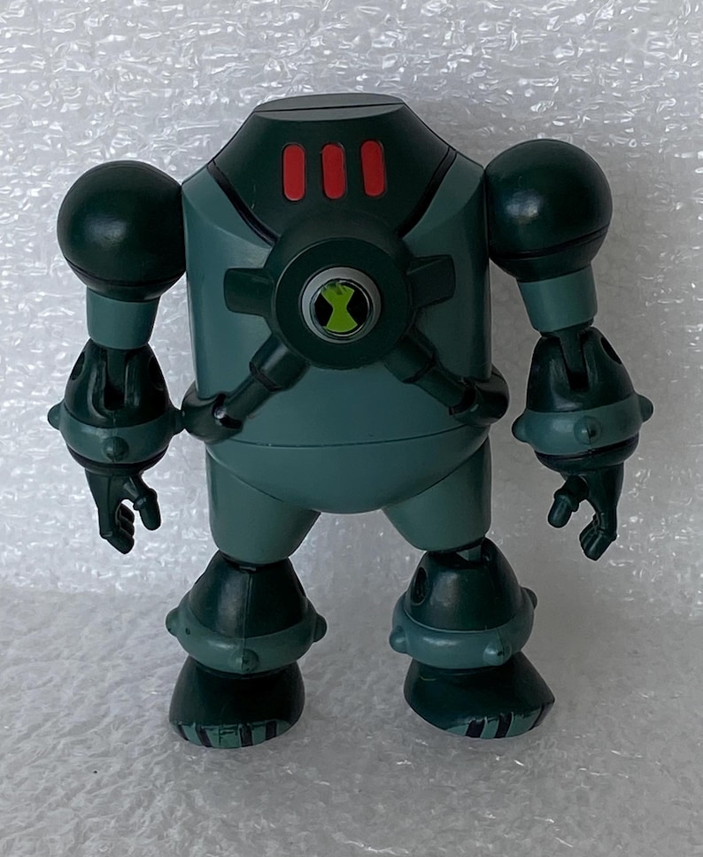May include: A teal robot action figure with a rounded head and three red rectangles. It features large spherical shoulders, articulated limbs, and a green and black emblem on its chest. The figure is a toy.