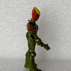 May include: A green and olive-colored action figure with a yellow and red head, featuring articulated joints at the shoulders, elbows, hips, and knees. The figure has black accents on the torso and red spikes on the back and shoulders.