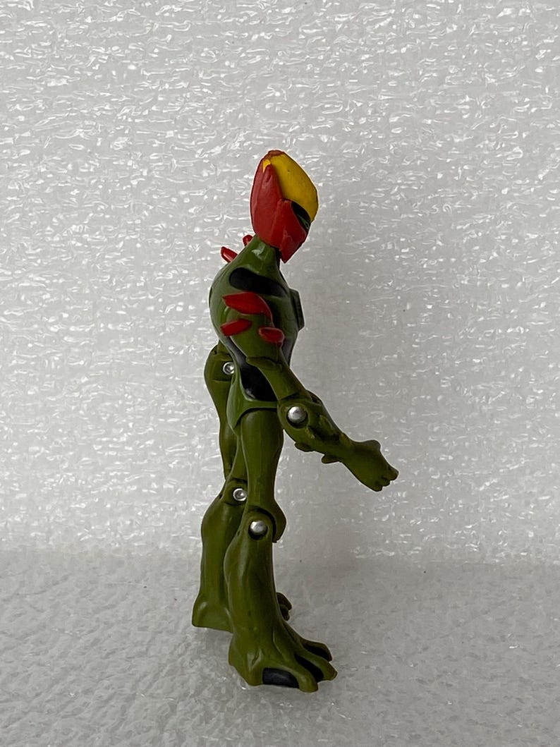 May include: A green and olive-colored action figure with a yellow and red head, featuring articulated joints at the shoulders, elbows, hips, and knees. The figure has black accents on the torso and red spikes on the back and shoulders.