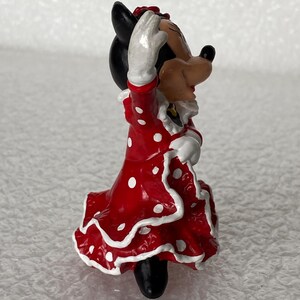 May include: A small figurine of a cartoon mouse character wearing a red dress with white polka dots and white ruffled trim. The character has black ears, a tan face, and white gloves, with one arm raised in a waving gesture while standing upright.