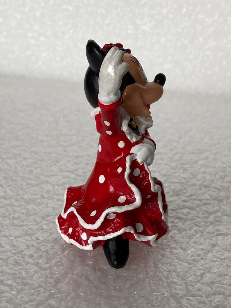 May include: A small figurine of a cartoon mouse character wearing a red dress with white polka dots and white ruffled trim. The character has black ears, a tan face, and white gloves, with one arm raised in a waving gesture while standing upright.
