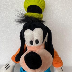 Może przedstawiać: Pluszowa zabawka przedstawiająca postać Goofy'ego z wysokim limonkowym kapeluszem z czarnym paskiem, długimi czarnymi uszami, brzoskwiniowym pyskiem z czarnym nosem, pomarańczową koszulką z długim rękawem, niebieskimi spodniami i białymi rękawiczkami.