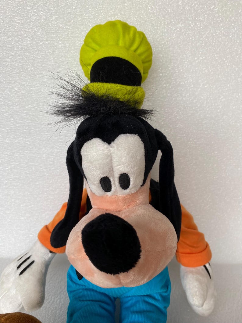 Może przedstawiać: Pluszowa zabawka przedstawiająca postać Goofy'ego z wysokim limonkowym kapeluszem z czarnym paskiem, długimi czarnymi uszami, brzoskwiniowym pyskiem z czarnym nosem, pomarańczową koszulką z długim rękawem, niebieskimi spodniami i białymi rękawiczkami.