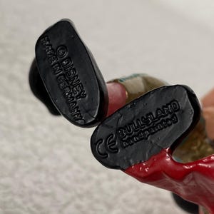 May include: The black soles of a figurine's feet feature molded text. One sole reads Disney and Made in Germany. The other sole displays the CE mark, the brand name Bullyland, and the text handpainted. The figurine has red and tan painted details.