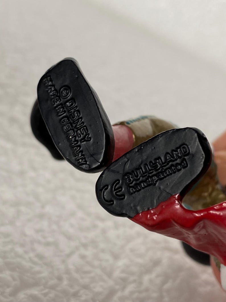 May include: The black soles of a figurine's feet feature molded text. One sole reads Disney and Made in Germany. The other sole displays the CE mark, the brand name Bullyland, and the text handpainted. The figurine has red and tan painted details.