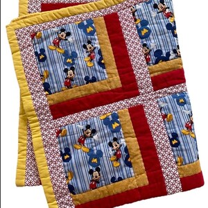 Pode incluir: Um edredom feito &agrave; m&atilde;o com um padr&atilde;o repetido do Mickey Mouse em um fundo listrado azul e branco. O edredom tem bordas vermelhas, douradas e amarelas, com um padr&atilde;o geom&eacute;trico nas bordas. O edredom est&aacute; dobrado.