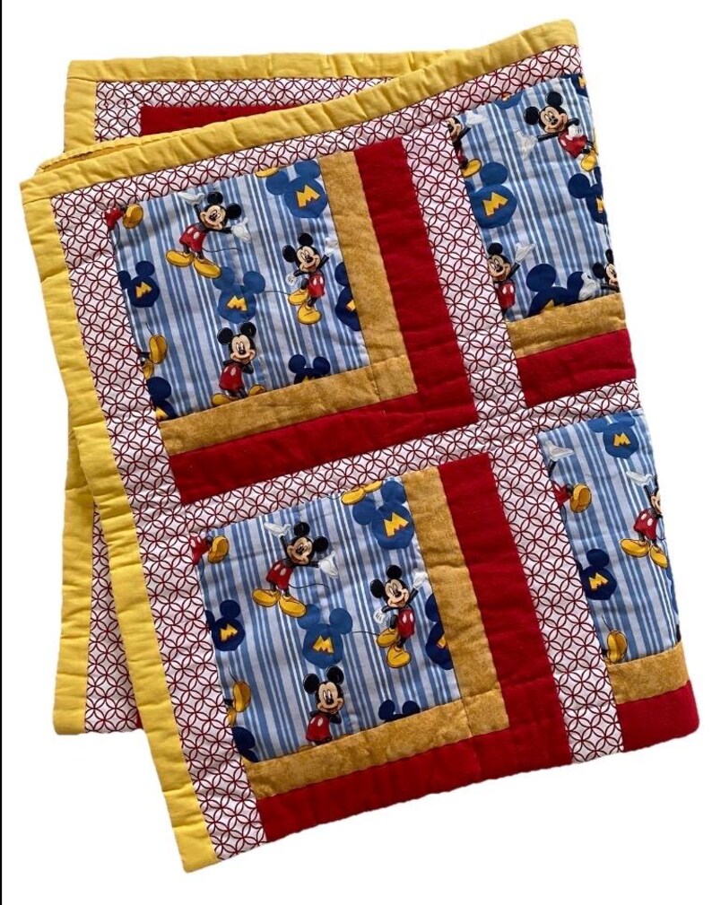 Pode incluir: Um edredom feito &agrave; m&atilde;o com um padr&atilde;o repetido do Mickey Mouse em um fundo listrado azul e branco. O edredom tem bordas vermelhas, douradas e amarelas, com um padr&atilde;o geom&eacute;trico nas bordas. O edredom est&aacute; dobrado.