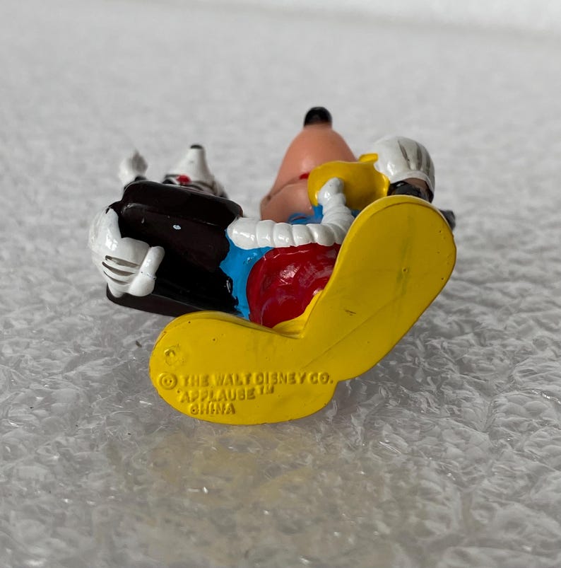 May include: A small Goofy figurine featuring a yellow shoe with the embossed text THE WALT DISNEY CO. APPLAUSE TM CHINA. The character wears a blue shirt, red pants, and white gloves, and is holding a black briefcase. The figure is made of plastic.