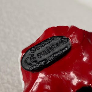 May include: A black oval base on a red and white object with the embossed text reading &copy; DISNEY, BULLYLAND, CE, and MADE IN GERMANY. The base is textured and features clear, raised lettering against a smooth, dark surface on the underside of the item.