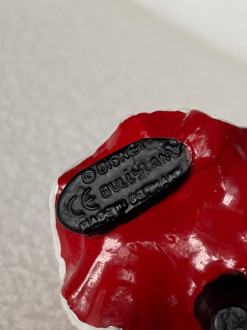 May include: A black oval base on a red and white object with the embossed text reading &copy; DISNEY, BULLYLAND, CE, and MADE IN GERMANY. The base is textured and features clear, raised lettering against a smooth, dark surface on the underside of the item.