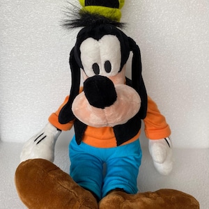 Może przedstawiać: Pluszowa zabawka przedstawiająca postać Goofy'ego, z wysokim limonkowym kapeluszem, długimi czarnymi uszami, czarną kamizelką na pomarańczowej koszulce z długim rękawem, jasnoniebieskimi spodniami i dużymi brązowymi butami oraz białymi rękawiczkami.