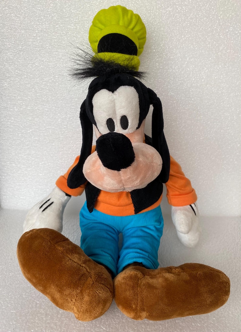 Może przedstawiać: Pluszowa zabawka przedstawiająca postać Goofy'ego, z wysokim limonkowym kapeluszem, długimi czarnymi uszami, czarną kamizelką na pomarańczowej koszulce z długim rękawem, jasnoniebieskimi spodniami i dużymi brązowymi butami oraz białymi rękawiczkami.