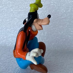 以下が含まれることがあります： ディズニーの漫画のキャラクター、グーフィーのカラフルなフィギュア。座った姿勢で、黒いベスト、オレンジ色のシャツ、青いズボン、茶色の靴、白い手袋、緑の帽子を着用しています。フィギュアは光沢のある素材で作られています。
