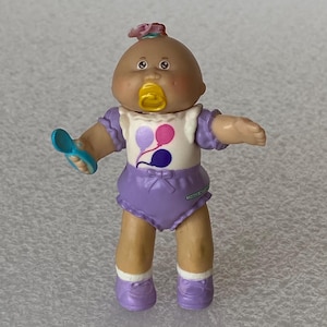 May include: A Cabbage Patch Kids doll with brown skin, wearing a purple diaper and shoes, and a white shirt with a balloon design. The doll has a yellow pacifier and a pink bow in its hair, holding a blue rattle.