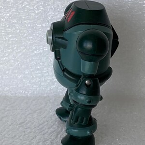 May include: A teal robot toy with a rounded body, spherical shoulder and elbow joints, and large feet. The robot has red accents on its head and silver details. The toy is standing on a white surface.