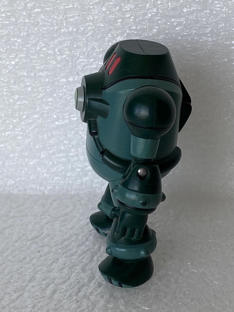 May include: A teal robot toy with a rounded body, spherical shoulder and elbow joints, and large feet. The robot has red accents on its head and silver details. The toy is standing on a white surface.