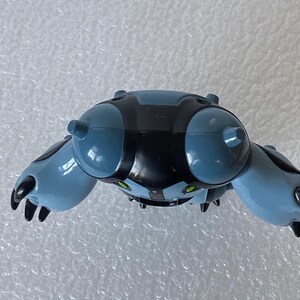 May include: A blue and black toy figure with a rounded body, two large, rounded appendages, and two smaller appendages. The figure has green eyes and black claws. The toy is made of plastic.