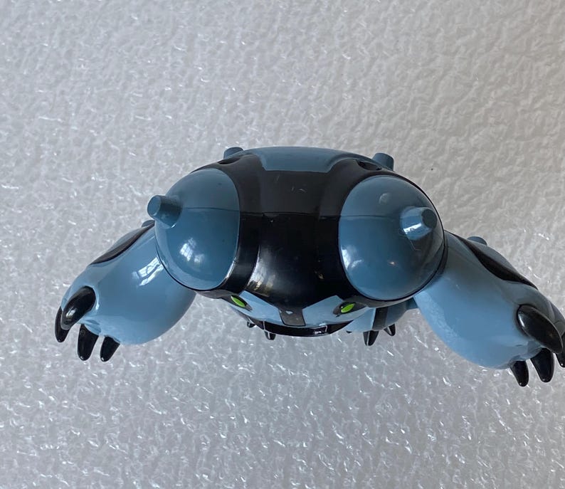 May include: A blue and black toy figure with a rounded body, two large, rounded appendages, and two smaller appendages. The figure has green eyes and black claws. The toy is made of plastic.