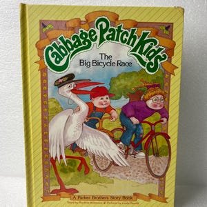 May include: A vintage children's book titled "Cabbage Patch Kids: The Big Bicycle Race." The cover features illustrations of children and a stork riding bicycles. The book has a yellow background with the title in green and the author's name at the bottom.