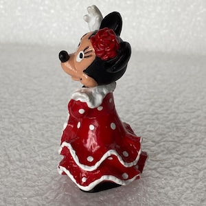 May include: A small figurine of a cartoon mouse character wearing a red dress with white polka dots and a white ruffled collar. The character has a red flower accessory on its head, black ears, and a white glove visible on one raised hand.