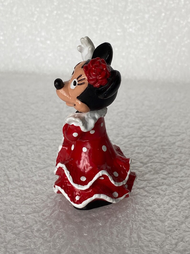 May include: A small figurine of a cartoon mouse character wearing a red dress with white polka dots and a white ruffled collar. The character has a red flower accessory on its head, black ears, and a white glove visible on one raised hand.