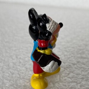 May include: A small figurine of a cartoon mouse character with black ears, a blue shirt, red shorts, and yellow shoes. The character is holding a dark brown object with both hands, featuring white gloves and a peach-colored face with a black nose.