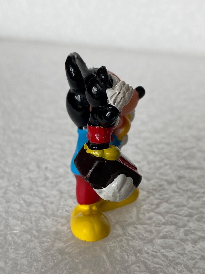 May include: A small figurine of a cartoon mouse character with black ears, a blue shirt, red shorts, and yellow shoes. The character is holding a dark brown object with both hands, featuring white gloves and a peach-colored face with a black nose.