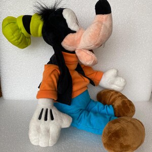 Może przedstawiać: Pluszowa zabawka przedstawiająca postać Goofy'ego z brzoskwiniowym pyszczkiem, czarnymi uszami i zielonym kapeluszem. Postać ma na sobie pomarańczową bluzkę z długim rękawem, czarną kamizelkę i jasnoniebieskie spodnie. Posiada białe rękawiczki i brązowe buty.