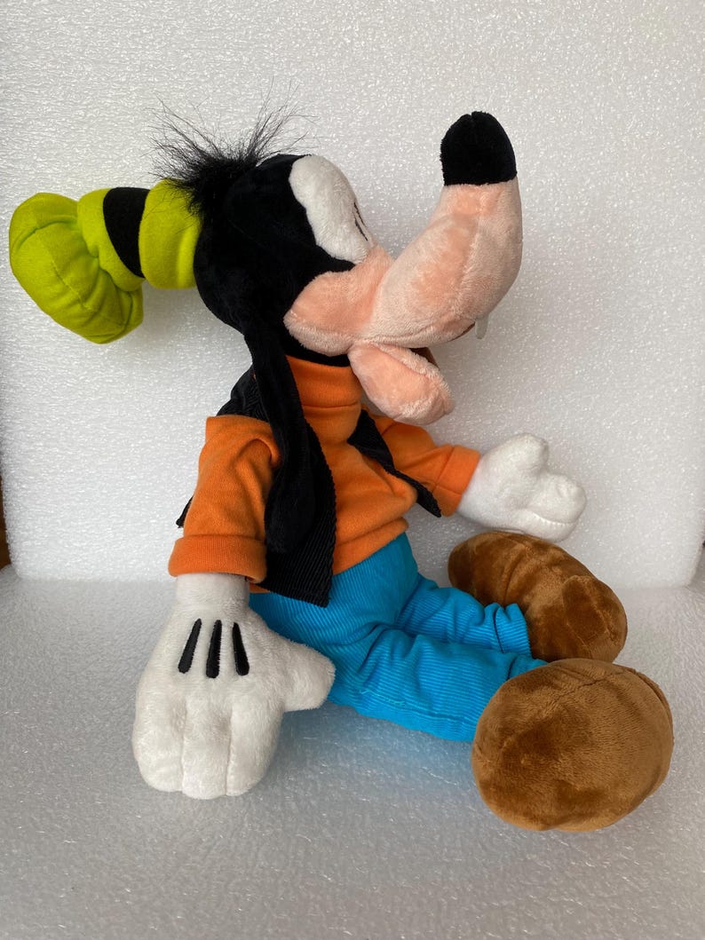 Może przedstawiać: Pluszowa zabawka przedstawiająca postać Goofy'ego z brzoskwiniowym pyszczkiem, czarnymi uszami i zielonym kapeluszem. Postać ma na sobie pomarańczową bluzkę z długim rękawem, czarną kamizelkę i jasnoniebieskie spodnie. Posiada białe rękawiczki i brązowe buty.