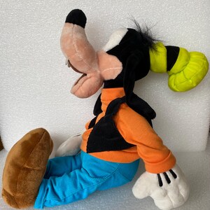 Może przedstawiać: Pluszowa zabawka przedstawiająca postać Goofy'ego, z brzoskwiniowym pyszczkiem, czarnymi uszami, zielonym kapeluszem, pomarańczową koszulką z długim rękawem, czarną kamizelką, niebieskimi spodniami sztruksowymi, białymi rękawiczkami i dużymi brązowymi butami.