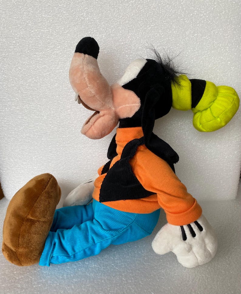 Może przedstawiać: Pluszowa zabawka przedstawiająca postać Goofy'ego, z brzoskwiniowym pyszczkiem, czarnymi uszami, zielonym kapeluszem, pomarańczową koszulką z długim rękawem, czarną kamizelką, niebieskimi spodniami sztruksowymi, białymi rękawiczkami i dużymi brązowymi butami.