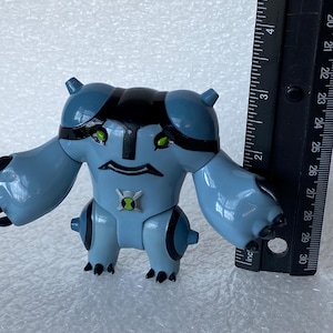 May include: A light blue action figure of a cartoon character with black accents and green eyes. The figure has a symbol on its chest and is approximately 3 inches tall. A black ruler is next to the figure.