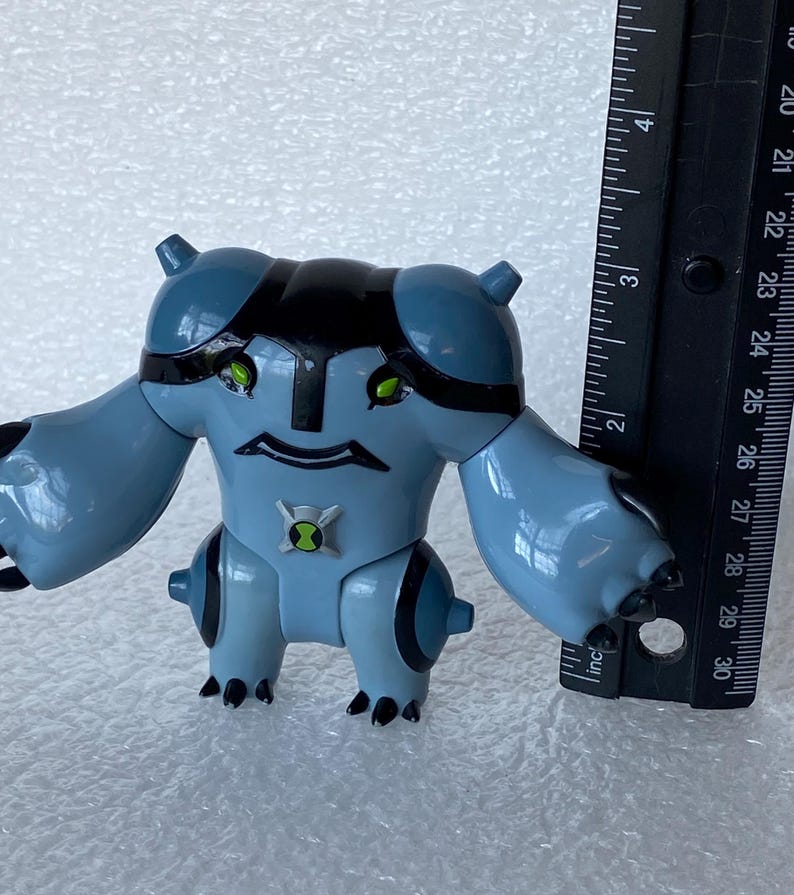 May include: A light blue action figure of a cartoon character with black accents and green eyes. The figure has a symbol on its chest and is approximately 3 inches tall. A black ruler is next to the figure.
