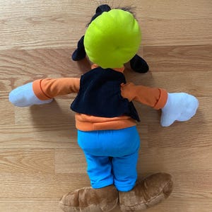 Może przedstawiać: Pluszowa zabawka przedstawiająca postać Goofy'ego, z jaskrawozielonym kapeluszem, pomarańczową koszulką z długim rękawem, czarną kamizelką sztruksową, jaskrawoniebieskimi spodniami, białymi rękawiczkami i brązowymi butami. Postać jest pokazana od tyłu.