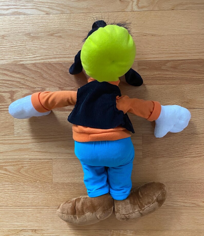 Może przedstawiać: Pluszowa zabawka przedstawiająca postać Goofy'ego, z jaskrawozielonym kapeluszem, pomarańczową koszulką z długim rękawem, czarną kamizelką sztruksową, jaskrawoniebieskimi spodniami, białymi rękawiczkami i brązowymi butami. Postać jest pokazana od tyłu.