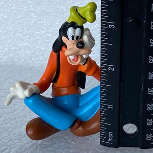 以下が含まれることがあります： ディズニーの漫画のキャラクター、グーフィーのカラフルなフィギュア。座ったポーズで、オレンジ色のシャツ、青いズボン、茶色の靴、黒いベスト、緑の帽子、白い手袋を着用しています。フィギュアの高さは約5cmです。