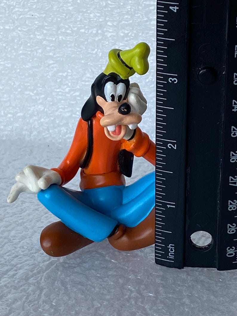 以下が含まれることがあります： ディズニーの漫画のキャラクター、グーフィーのカラフルなフィギュア。座ったポーズで、オレンジ色のシャツ、青いズボン、茶色の靴、黒いベスト、緑の帽子、白い手袋を着用しています。フィギュアの高さは約5cmです。
